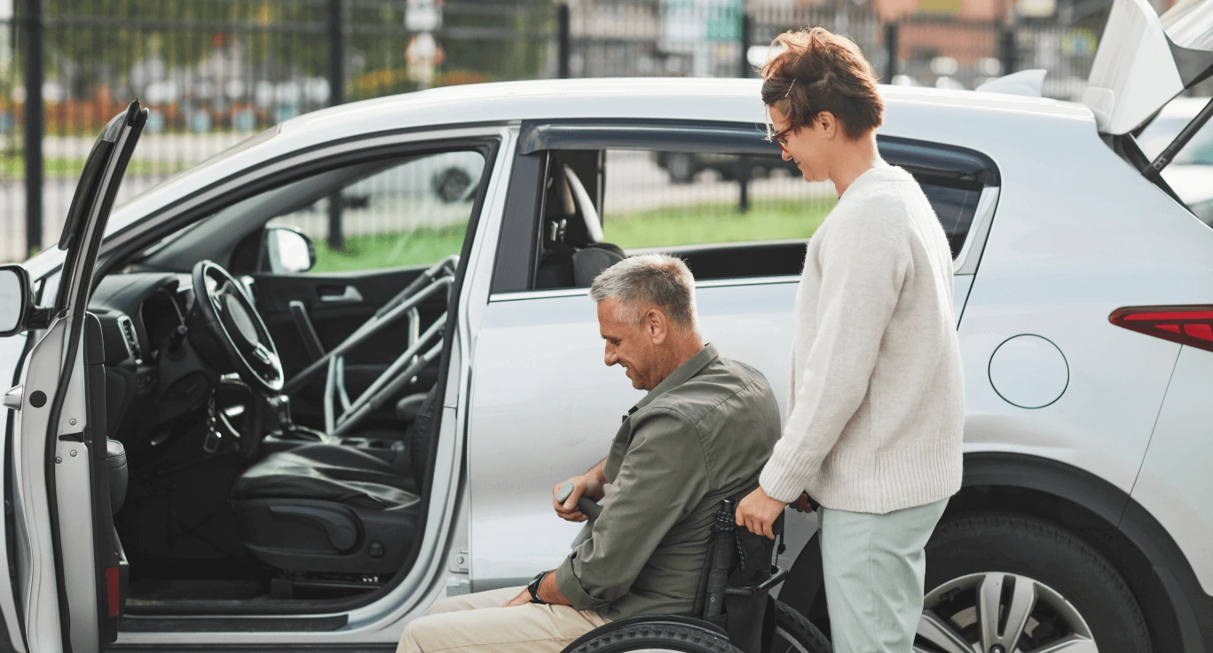 Woman helping a man move from his wheelchair into a white SUV