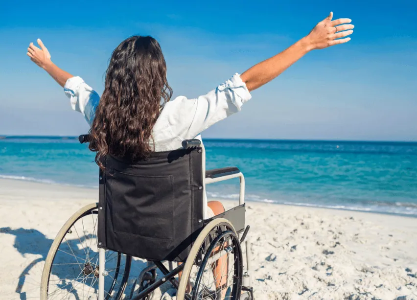 Woman in a wheelchair on a beach with her arms raised, facing the ocean